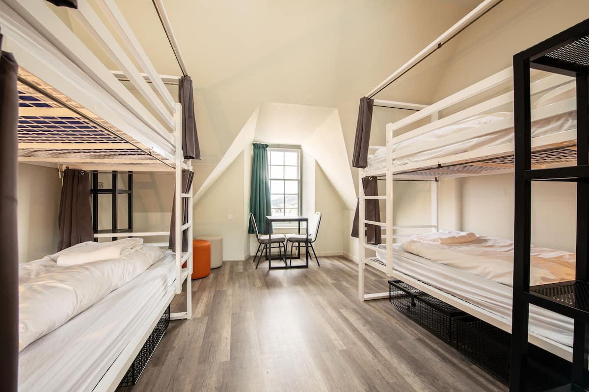 A dormitory room with bunk beds, a small table, and a window with curtains.