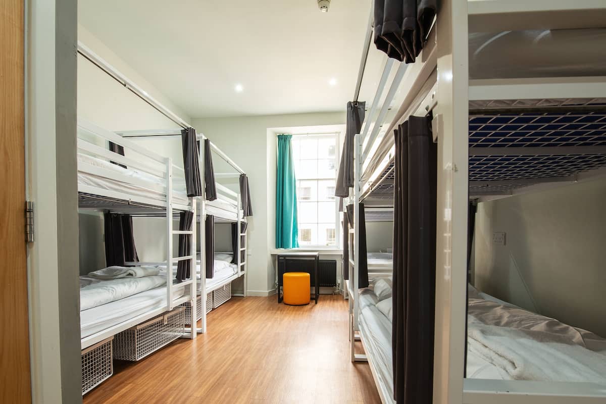 A dormitory room with bunk beds, a window with curtains, a radiator, and a small orange ottoman.