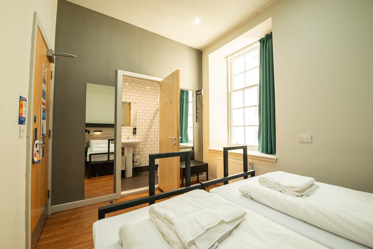 A hotel room with a wooden floor, a bed with white linens, a small table, a chair, a bathroom with a sink and mirror, and a window with curtains.