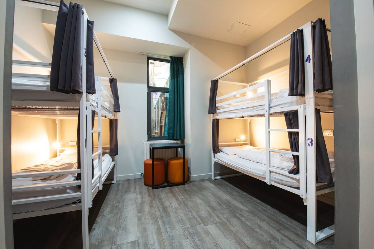 A dormitory room with bunk beds, a window with curtains, and a small table with two orange stools.