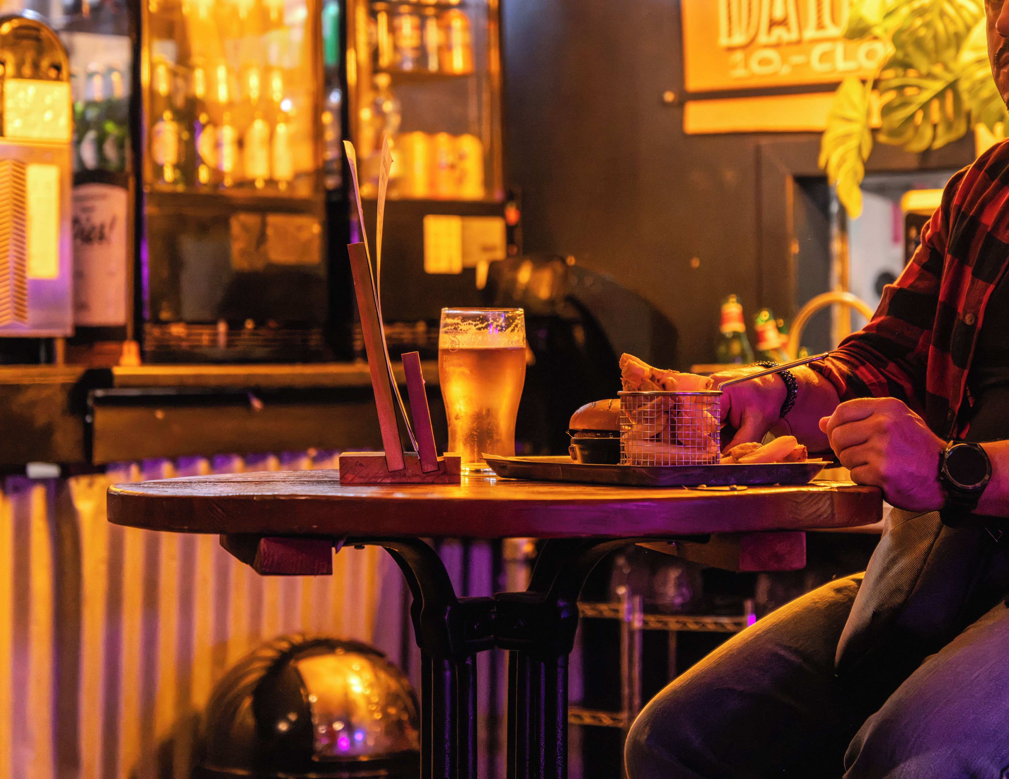 A bar setting with a person seated at a table, a glass of beer, and a basket of food items.