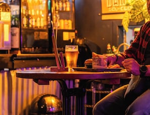A bar setting with a person seated at a table, a glass of beer, and a basket of food items. - a&o Hostel Edinburgh City (Edinburgh)