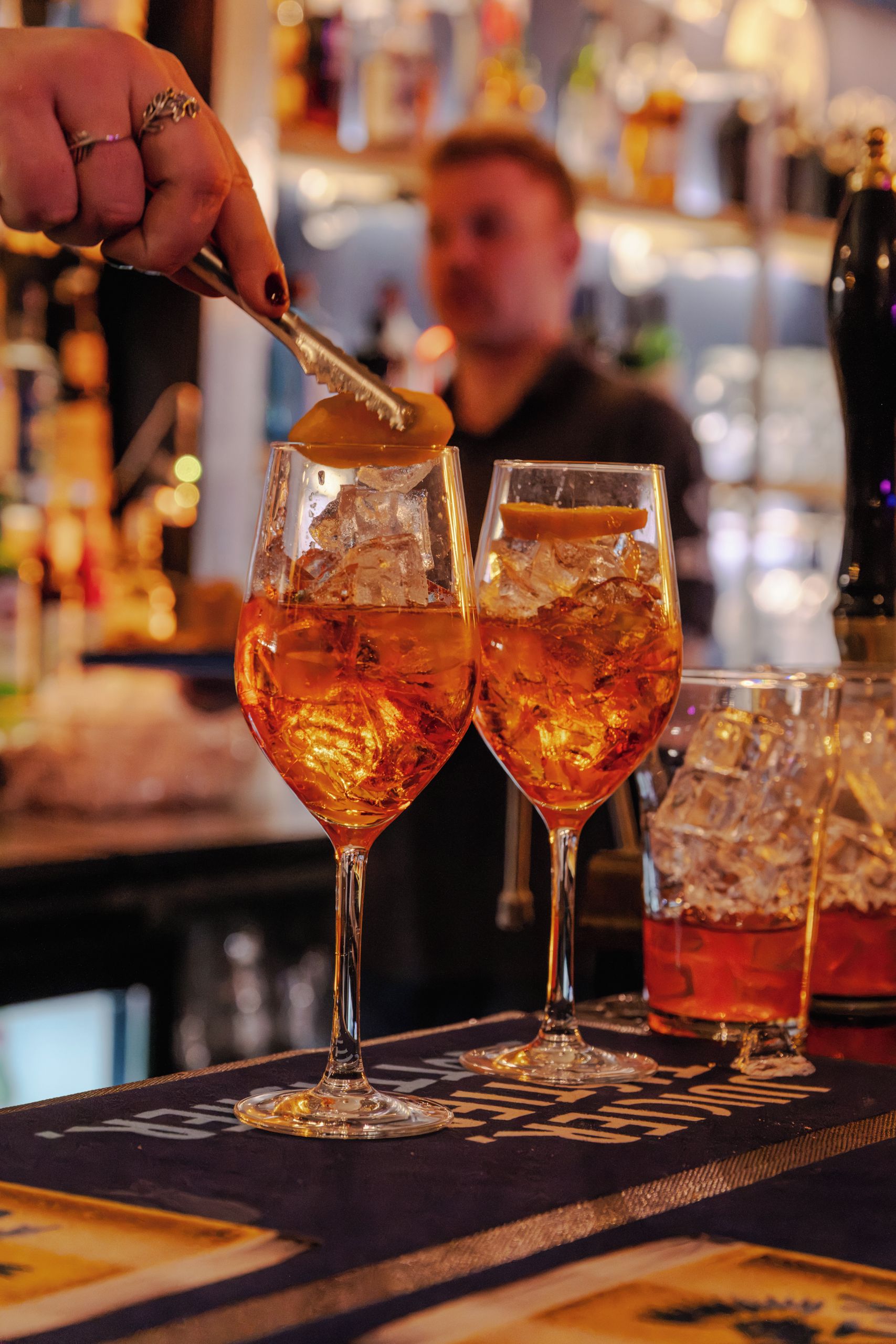 A bar setting with two glasses of amber-colored drink, ice, and a bar spoon.