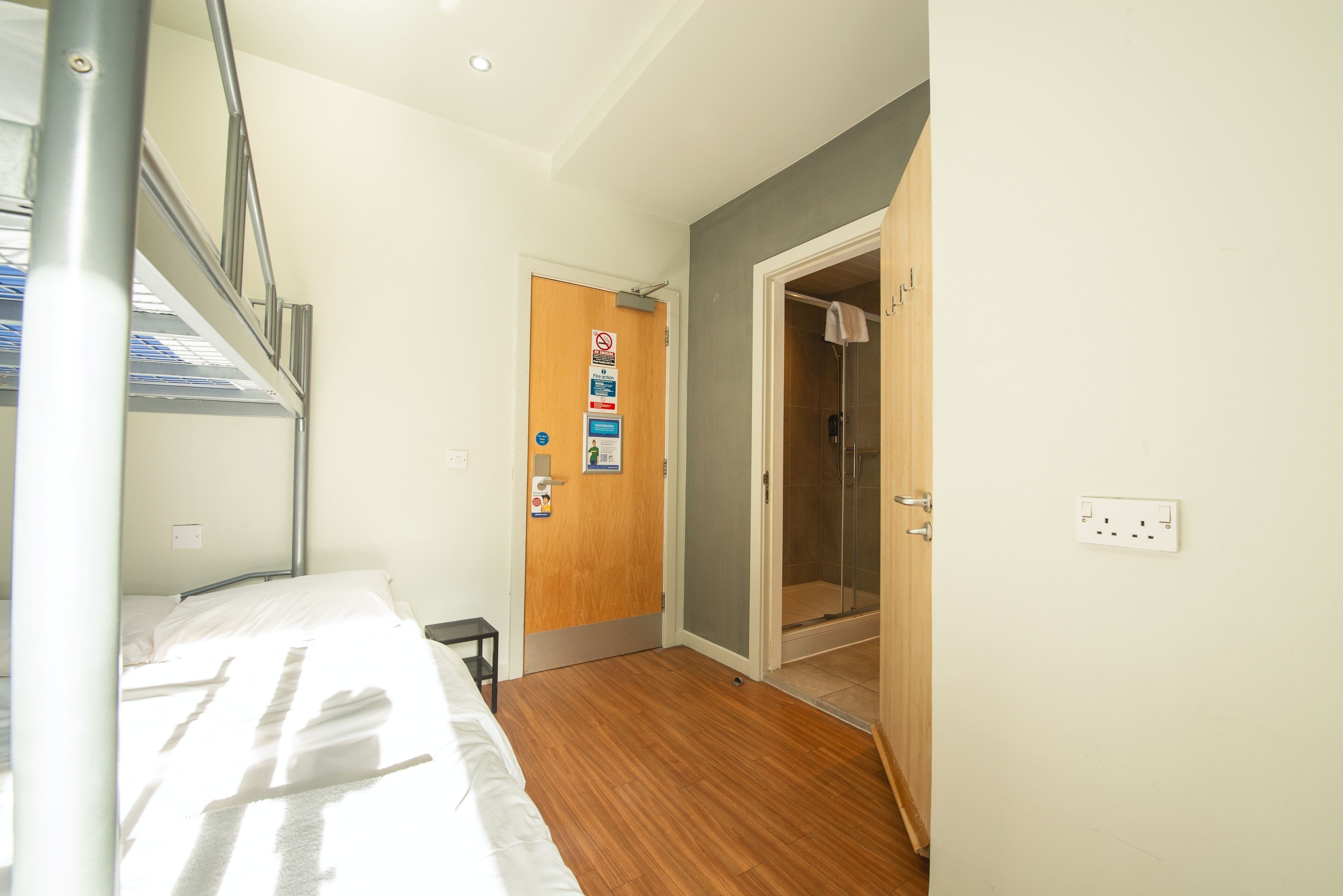 Habitación de hotel con cama, suelo de madera, zona de ducha y puerta con una toalla colgada.