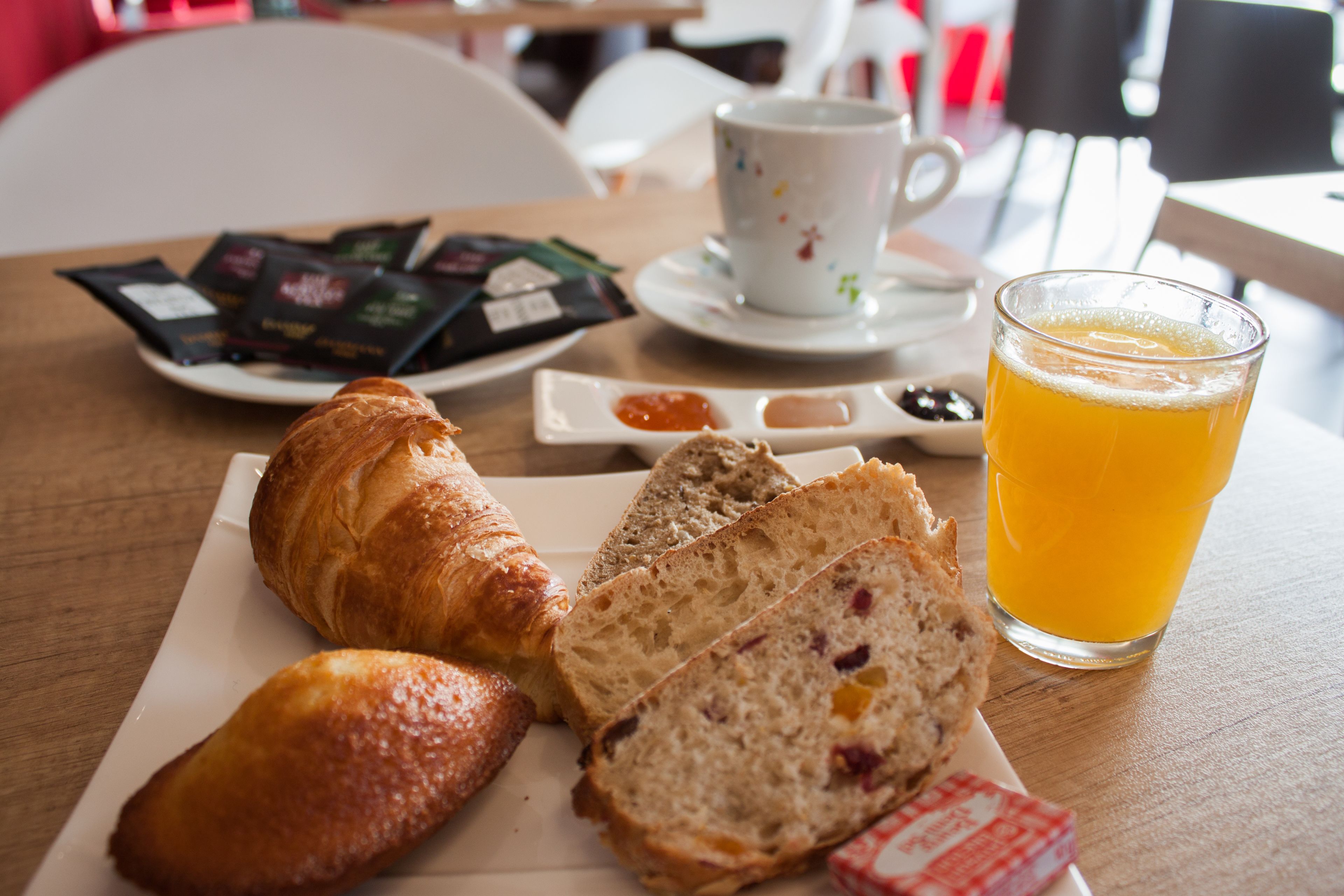 Mesa posta para o café da manhã, com croissants, pão, um copo de suco de laranja e sachês de chá e café.