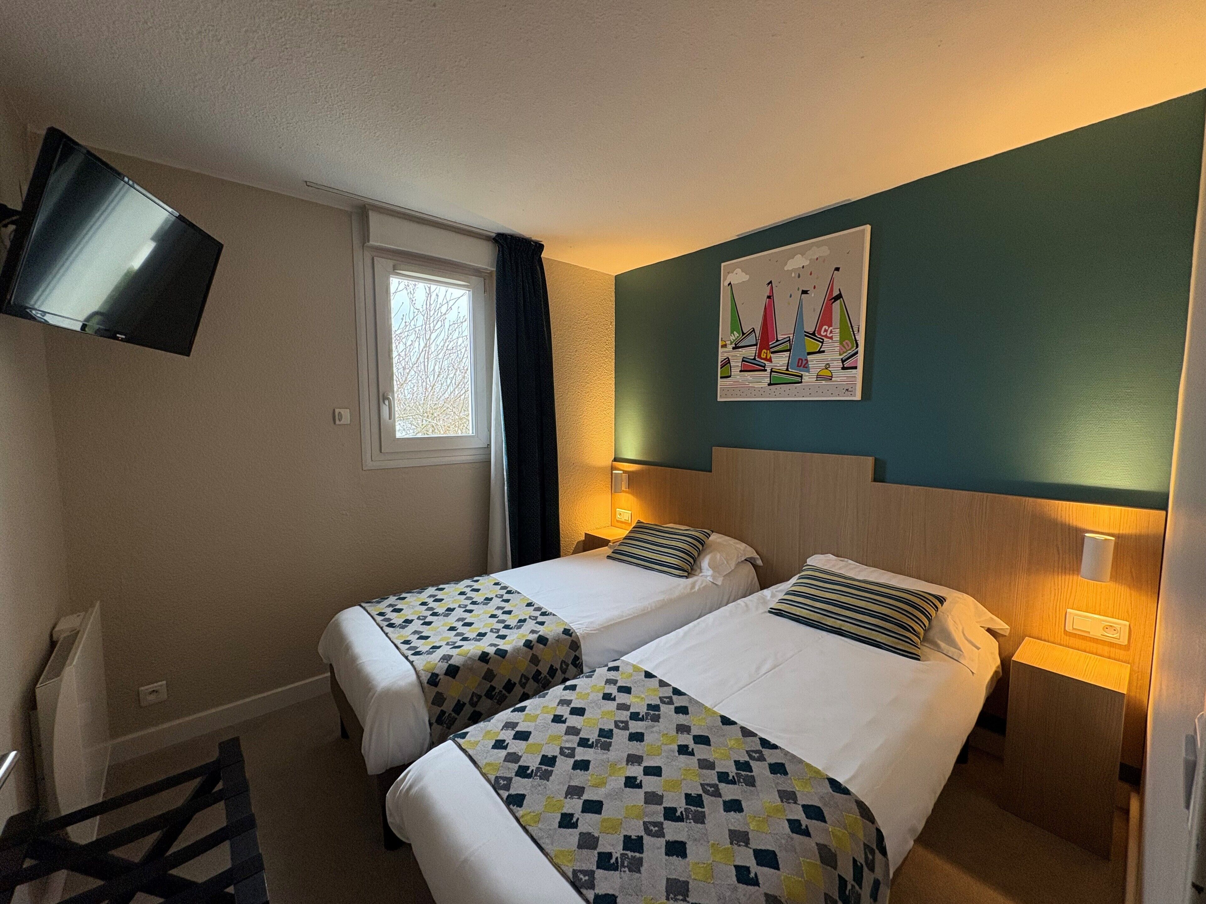 Habitación de hotel con dos camas, un televisor montado en la pared, una ventana con cortinas y un cuadro de veleros.