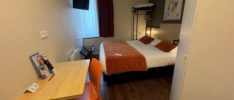 A hotel room with a bed, a desk with a chair, a window with curtains, a bedside table, and a wall-mounted TV.