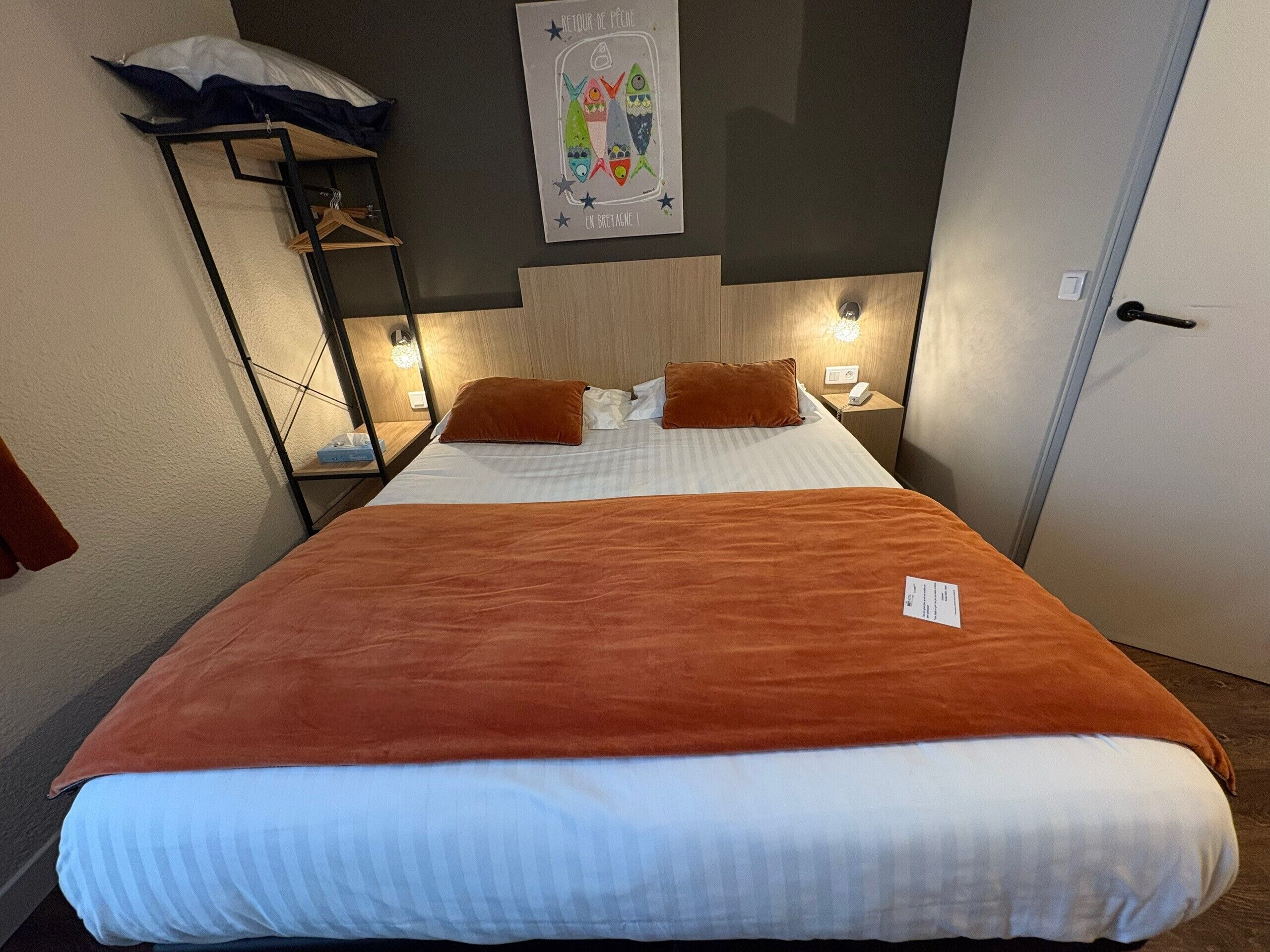 Una habitación de hotel con una cama, una mesita de noche, un estante en la pared y una puerta.