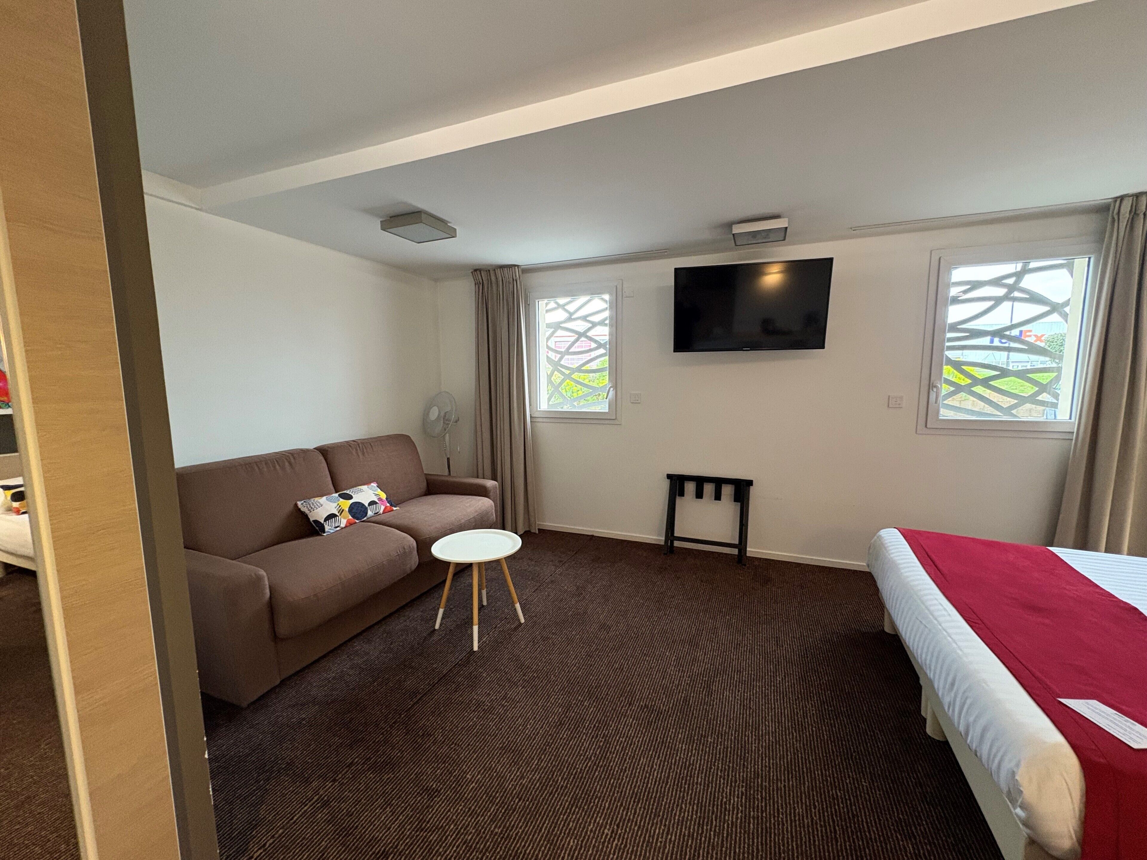 A hotel room with a brown sofa, a small white table, a bed with a red blanket, a TV on the wall, and two windows with curtains.