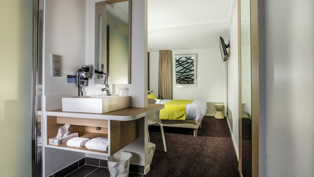 Habitación de hotel con baño, cama, mesita de noche, espejo y ventana con cortinas.