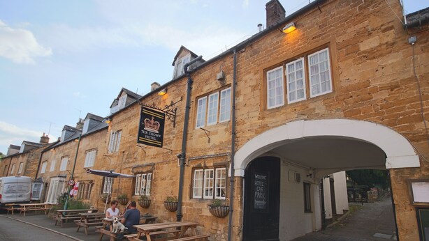 A stone building with a sign for "The Crown Hotel" and a "Welcome Hotel Car Park" sign. There are outdoor tables and benches, and a van parked on the street. - The Crown Hotel (Moreton-in-Marsh)