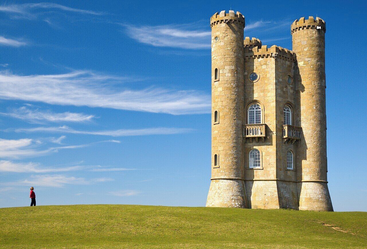 A large stone tower with multiple windows and a balcony, surrounded by a grassy hill under a clear sky.