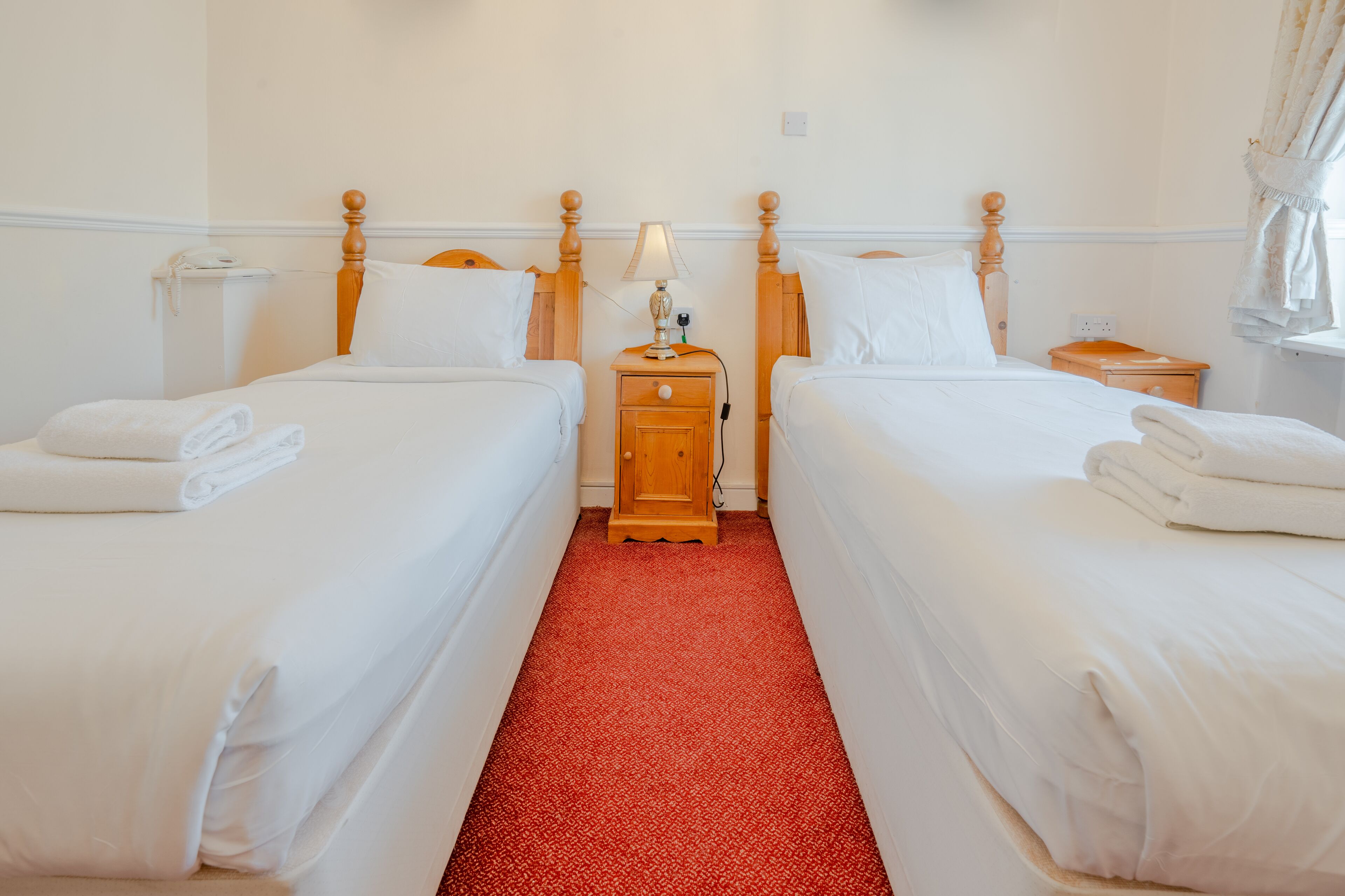 A hotel room with two beds, a red carpet, wooden nightstands, and a lamp.
