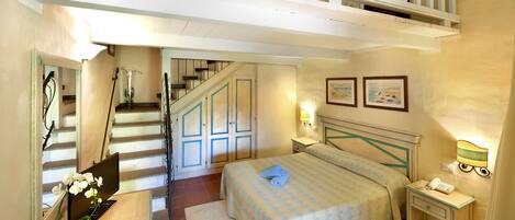 A bedroom with a bed, a desk with a computer and a vase of flowers, a nightstand with a lamp, and a staircase leading to an upper level.