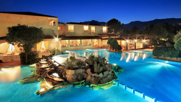 A swimming pool with a rock island in the center, surrounded by a building with multiple balconies and outdoor seating areas. - Colonna Hotel Du Golf (Olbia)