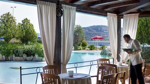 A poolside dining area with tables and chairs, a waiter serving a guest, and a view of a pool and landscape. - Colonna Hotel Du Golf (Olbia)