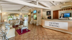 Eine Hotellobby mit einer Rezeption mit der Aufschrift "Colonna Country & Sporting". Es gibt eine hohe Decke mit freiliegenden Balken, eine Sitzecke mit Sofas und Tischen sowie einen großen Fernsehbildschirm.