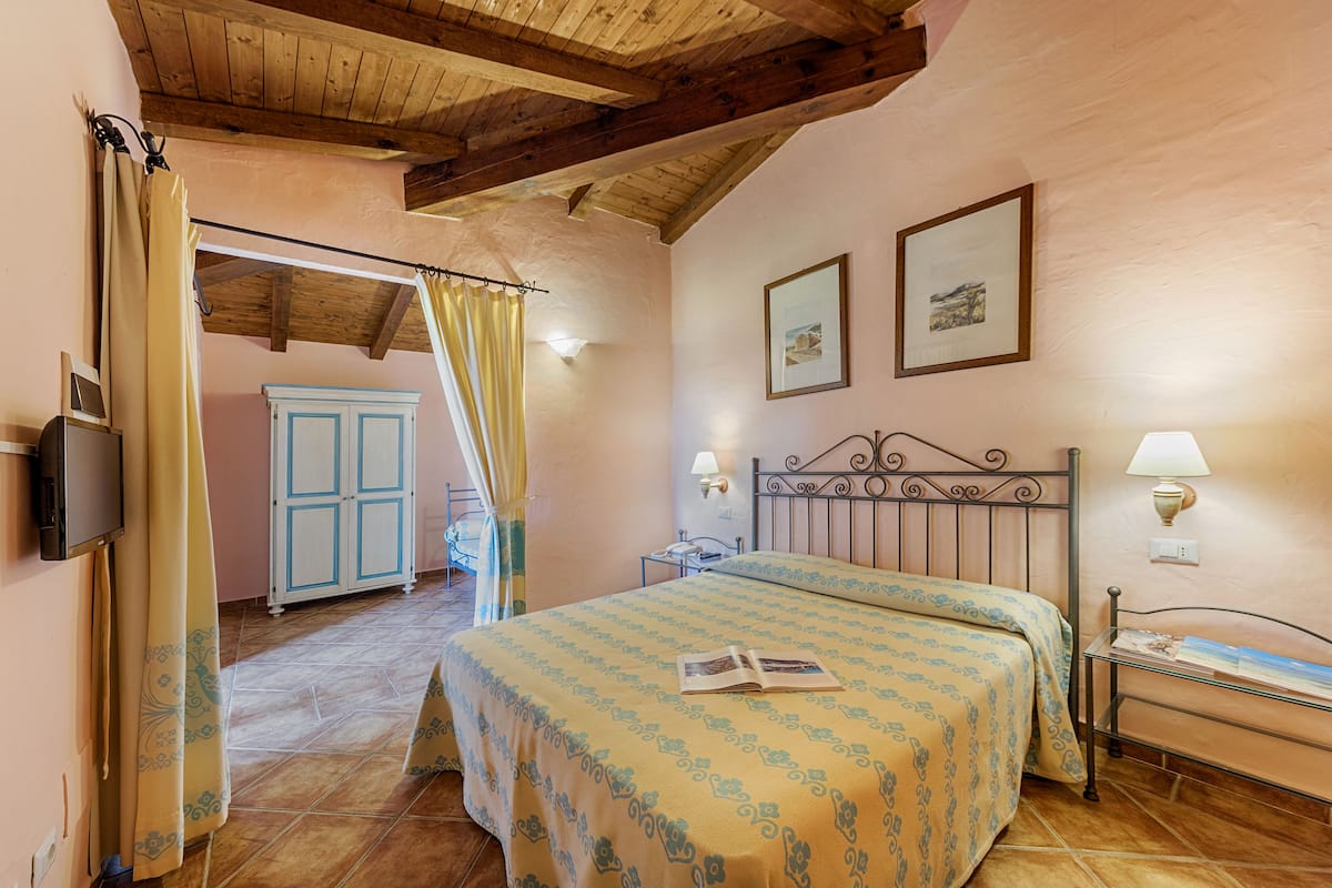 A bedroom with a bed, a nightstand, a lamp, a wardrobe, and a door leading to another room.