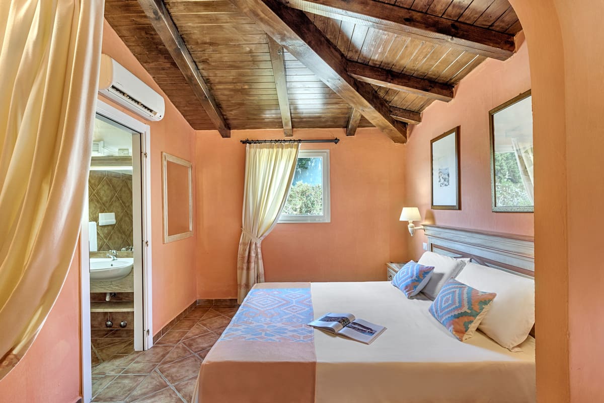 A bedroom with a bed, a window, an air conditioning unit, and a bathroom visible through an open door.