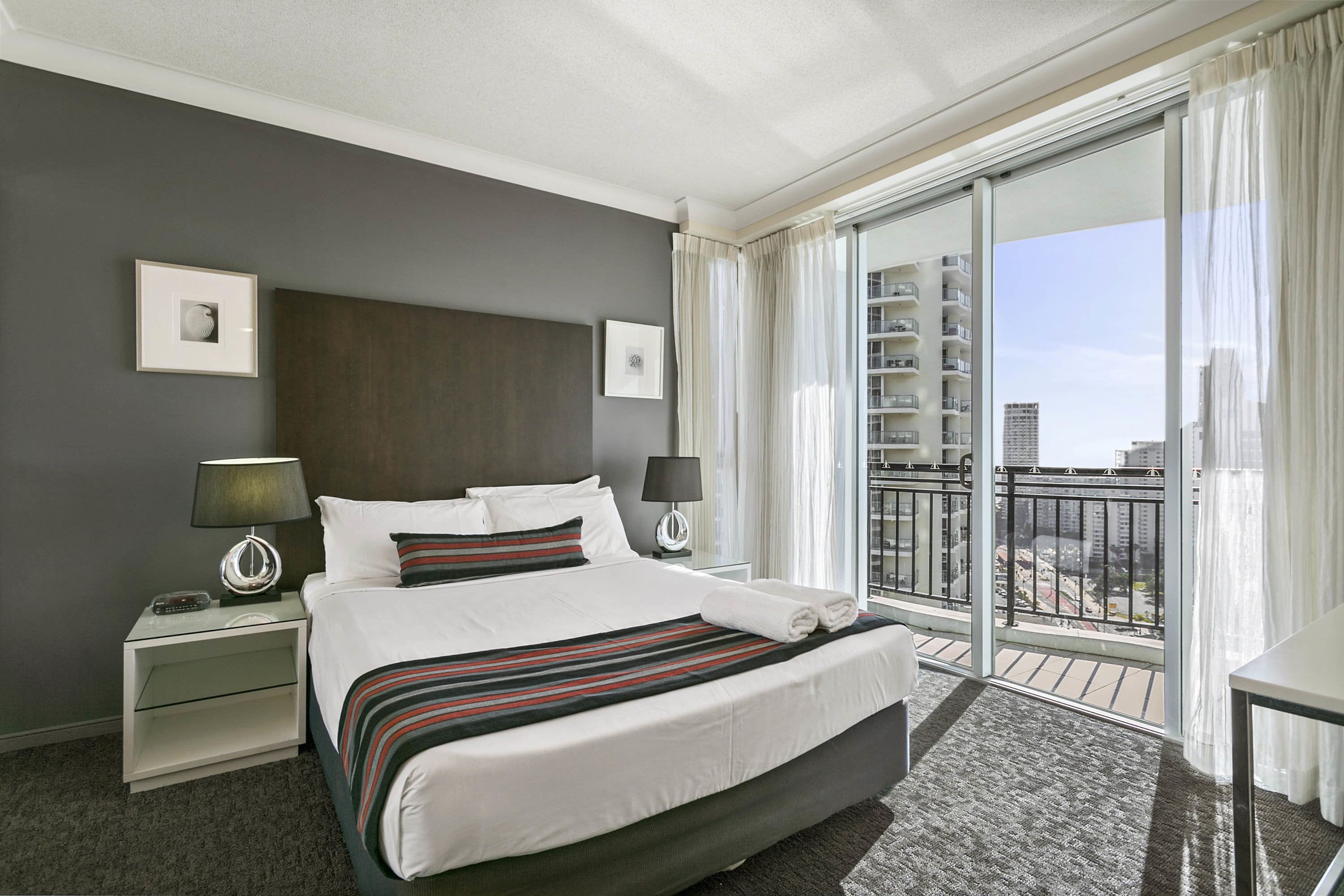 A hotel room with a large bed, two bedside tables with lamps, a view of a cityscape through a balcony.