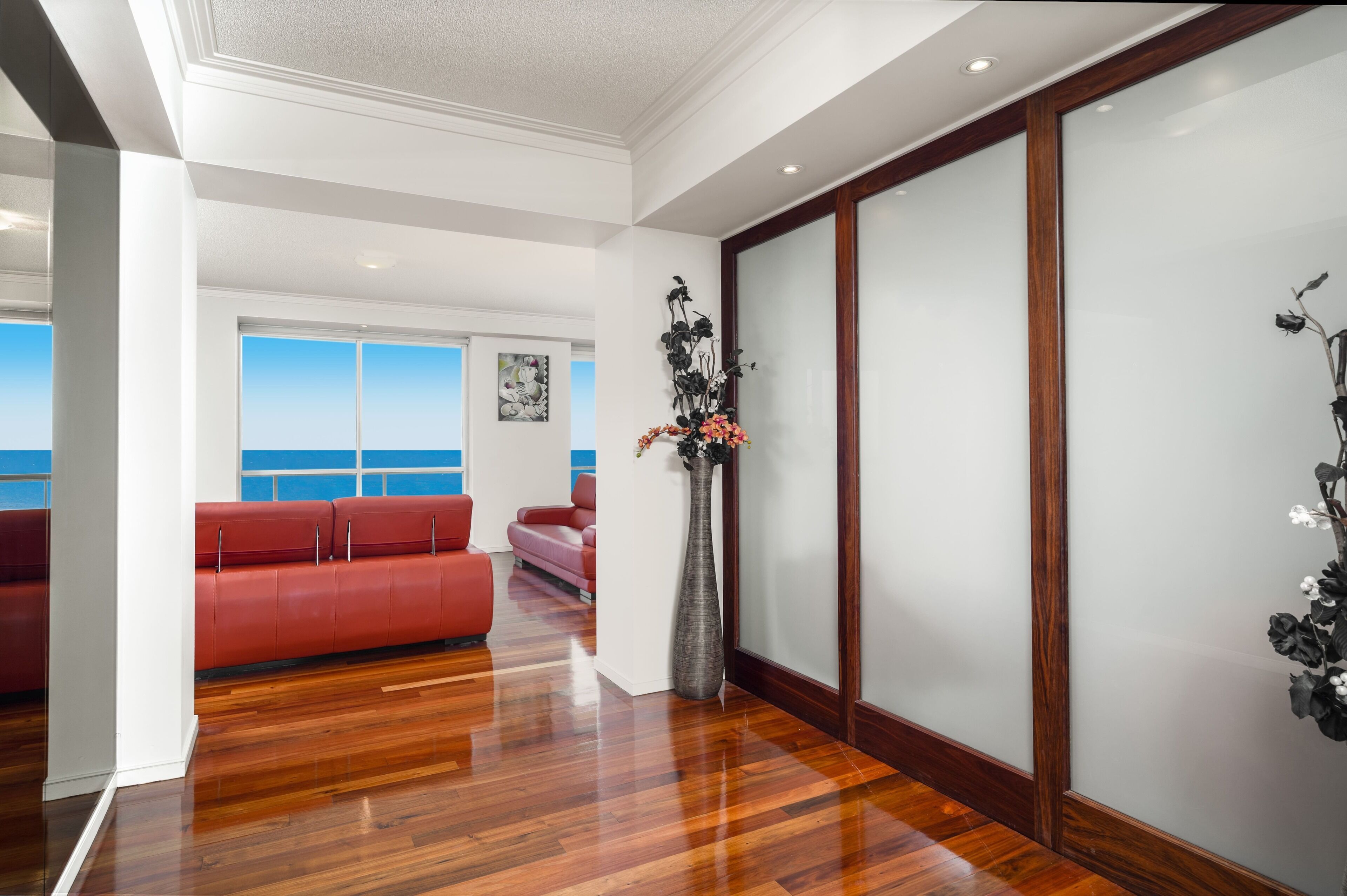 A modern interior with a wooden floor, red seating, a vase with flowers, and a view of the sea.