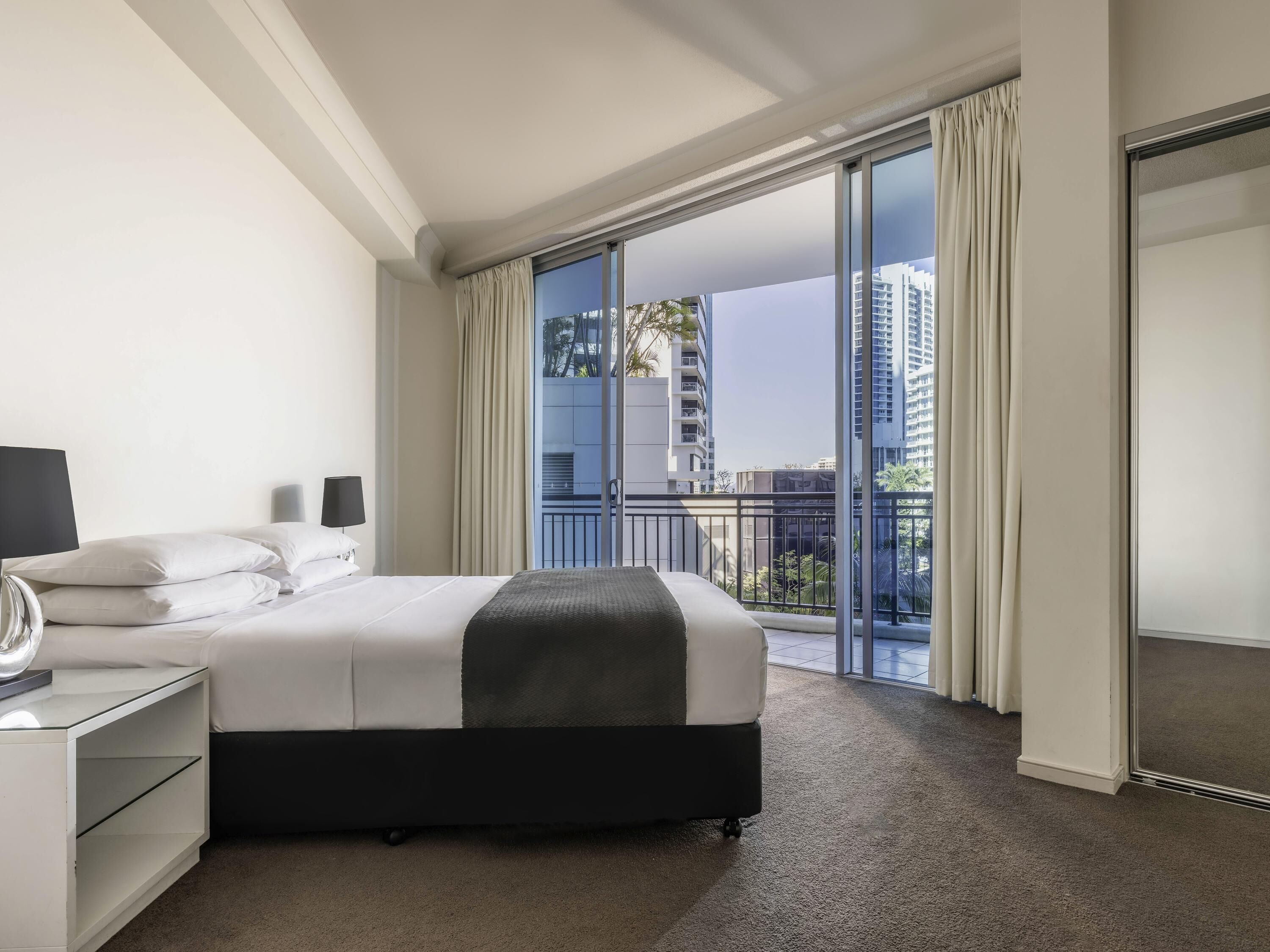A hotel room with a bed, a nightstand, a lamp, a large window with curtains, and a view of buildings.
