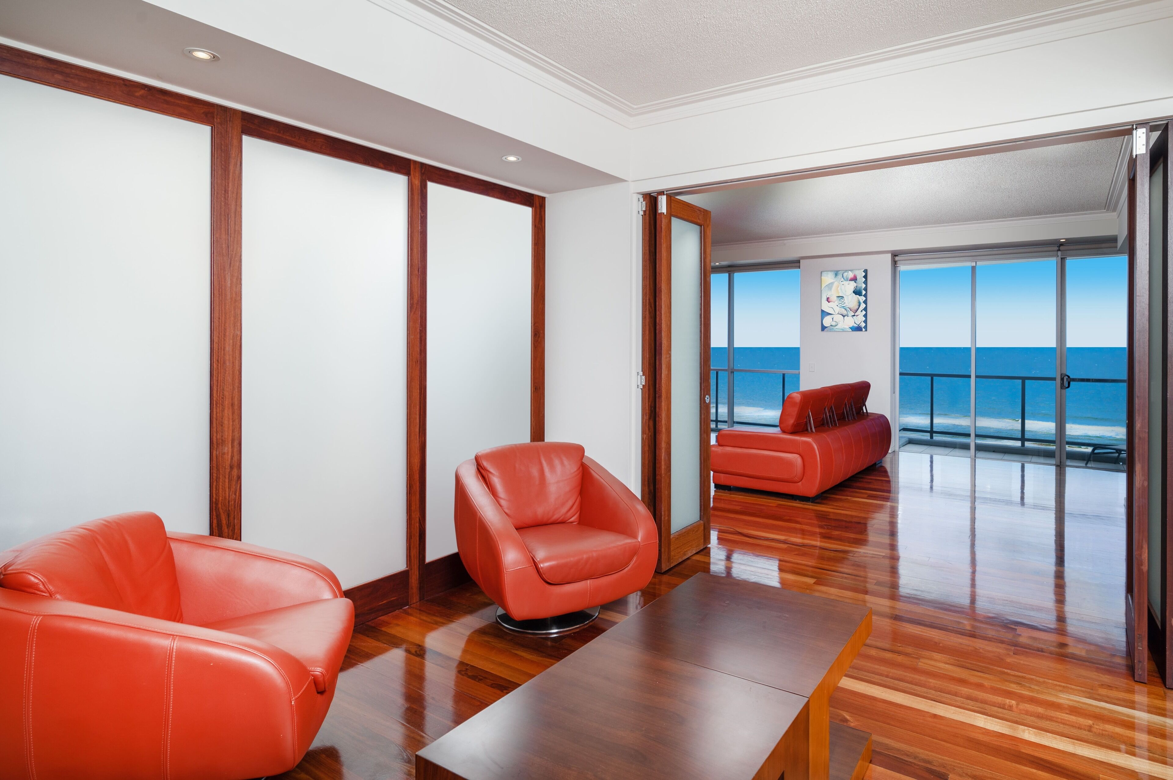 A modern living room with a wooden floor, a red leather armchair, and a view of the ocean through large glass doors.