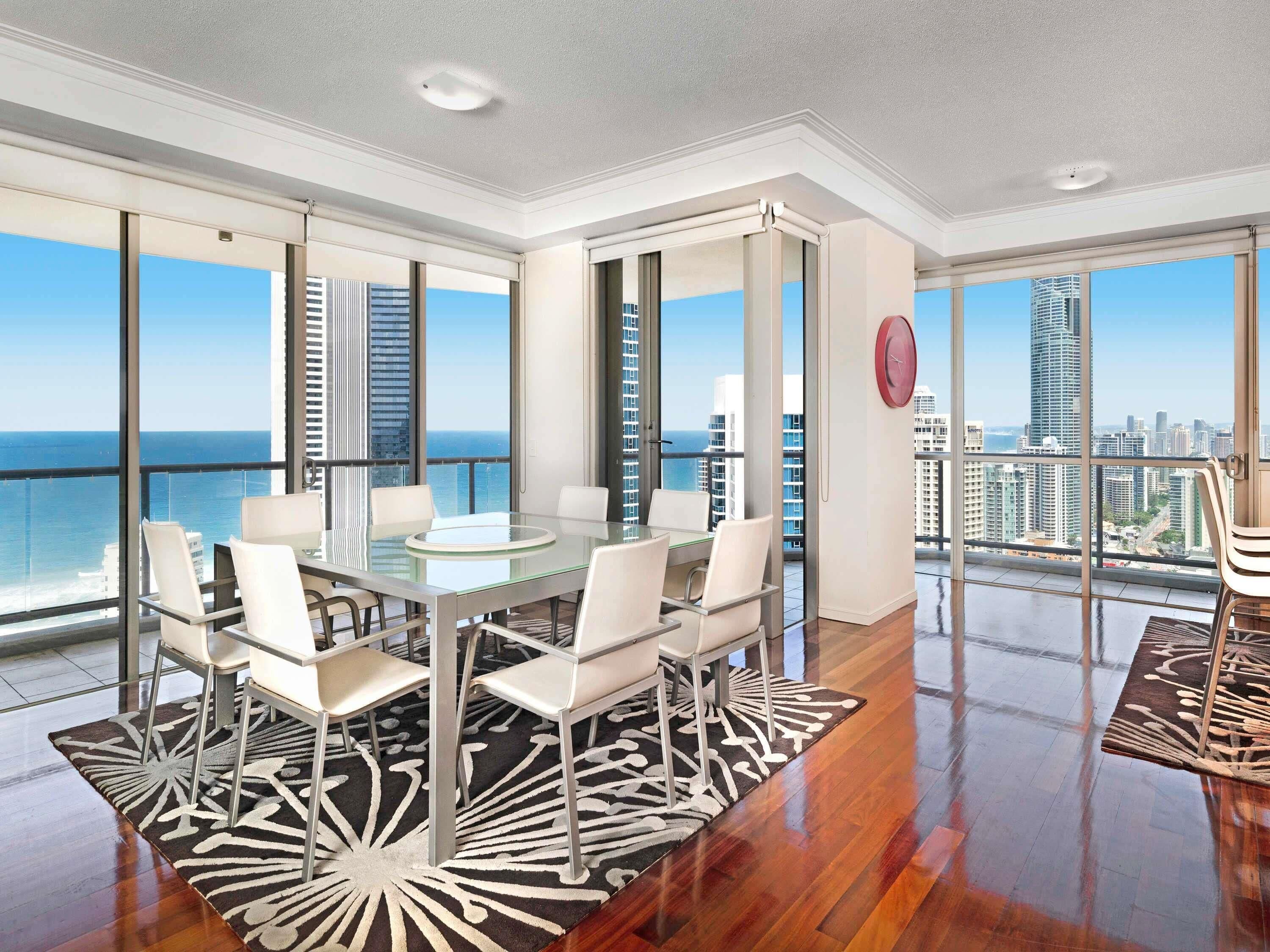 Um quarto espa çoso com mesa de vidro, cadeiras brancas e vista para o oceano e o horizonte da cidade.