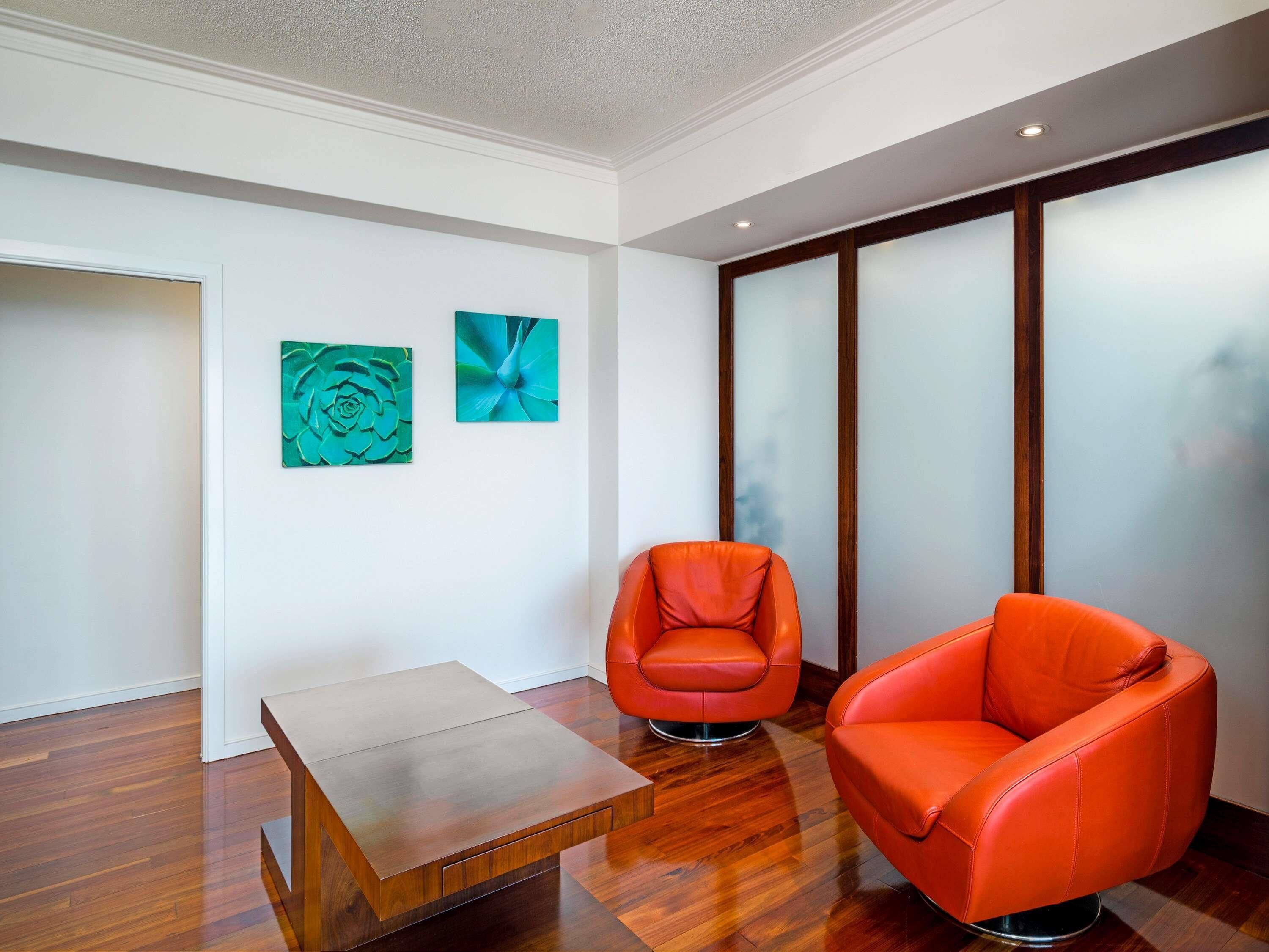 A modern interior with a wooden floor, two orange chairs, a small wooden table, and two abstract wall art pieces.