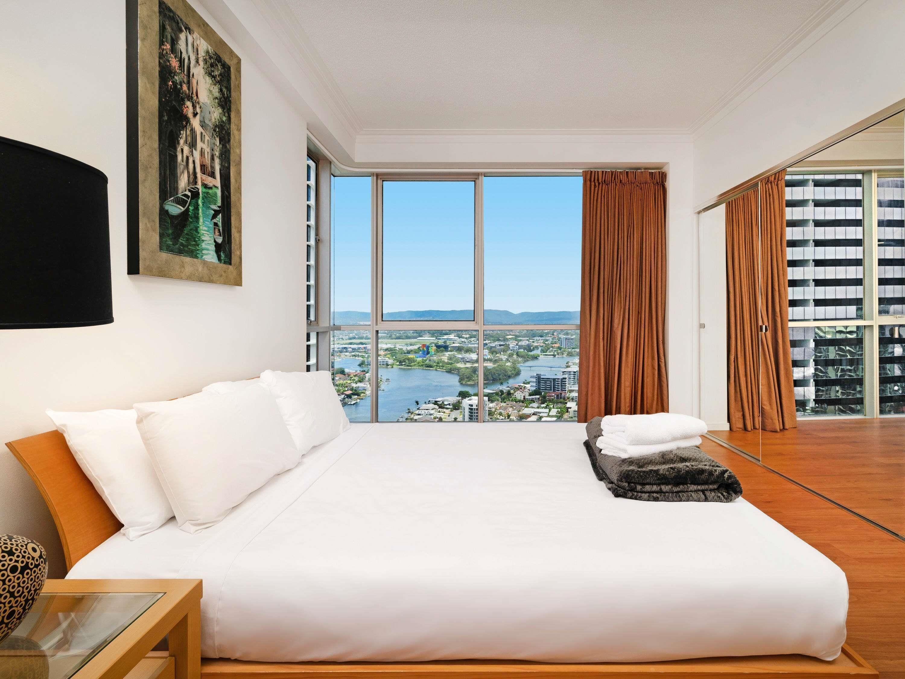 A bedroom with a large bed, a painting, a view of a cityscape, and a sliding glass door.