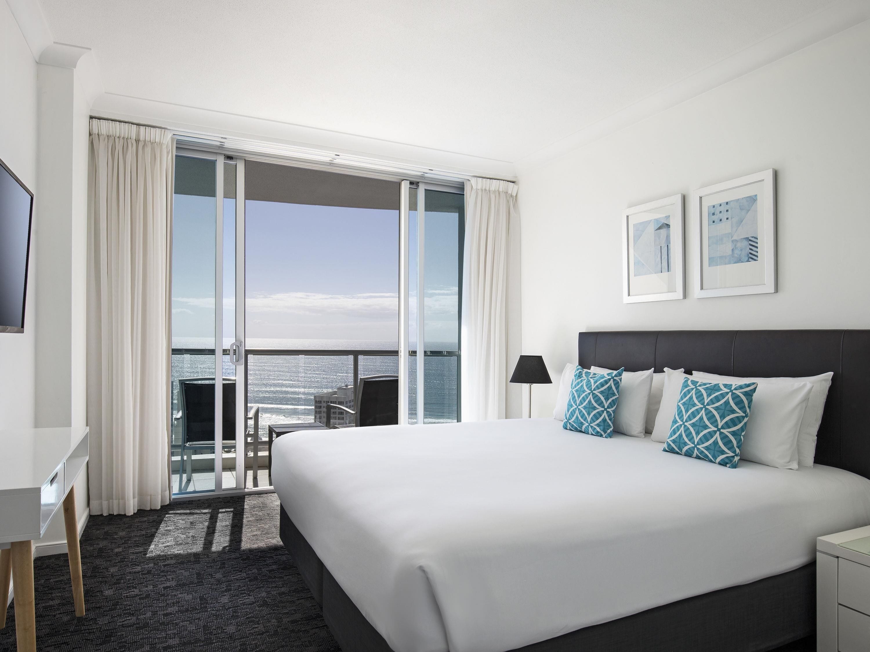 A hotel room with a large bed, a desk, a chair, a TV, and a balcony with a view of the ocean.