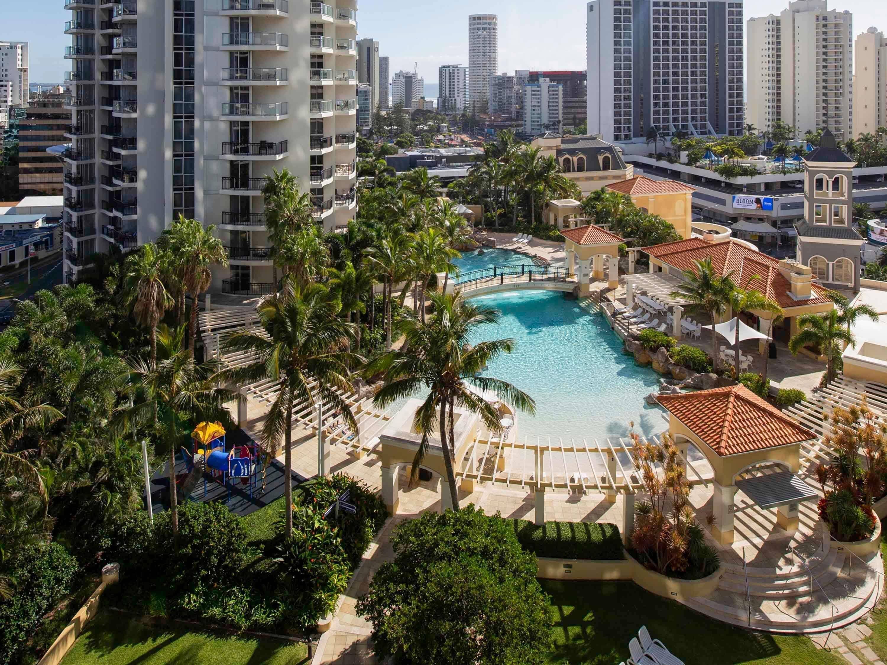 A resort with a central swimming pool, palm trees, and high-rise buildings in the background.