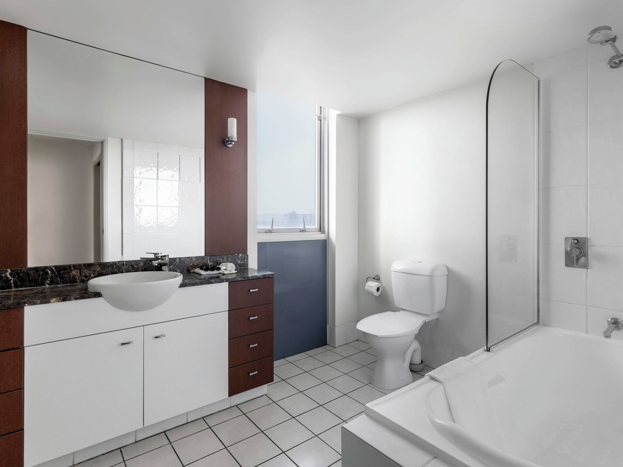 Un baño moderno con un lavamanos blanco, encimera de mármol oscuro, armarios blancos, inodoro blanco y ducha con mampara de cristal.