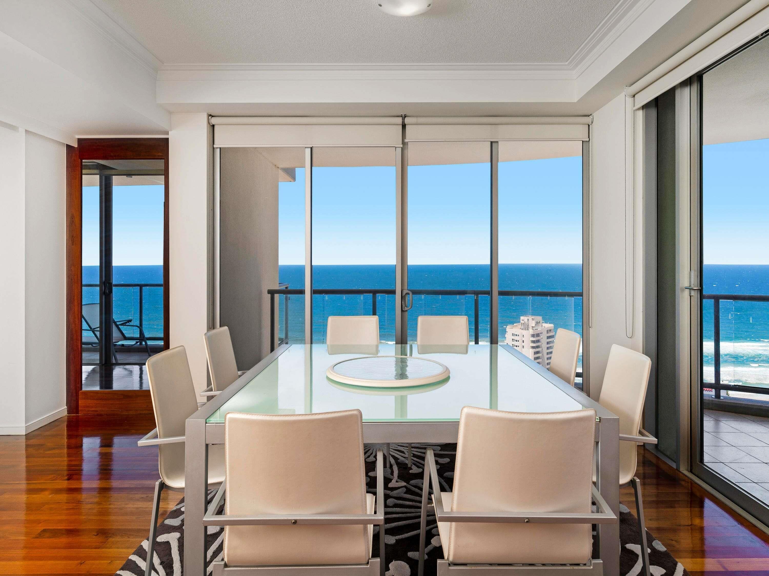 A modern dining area with a glass table and chairs, large windows with a view of the ocean, and a balcony with a railing.