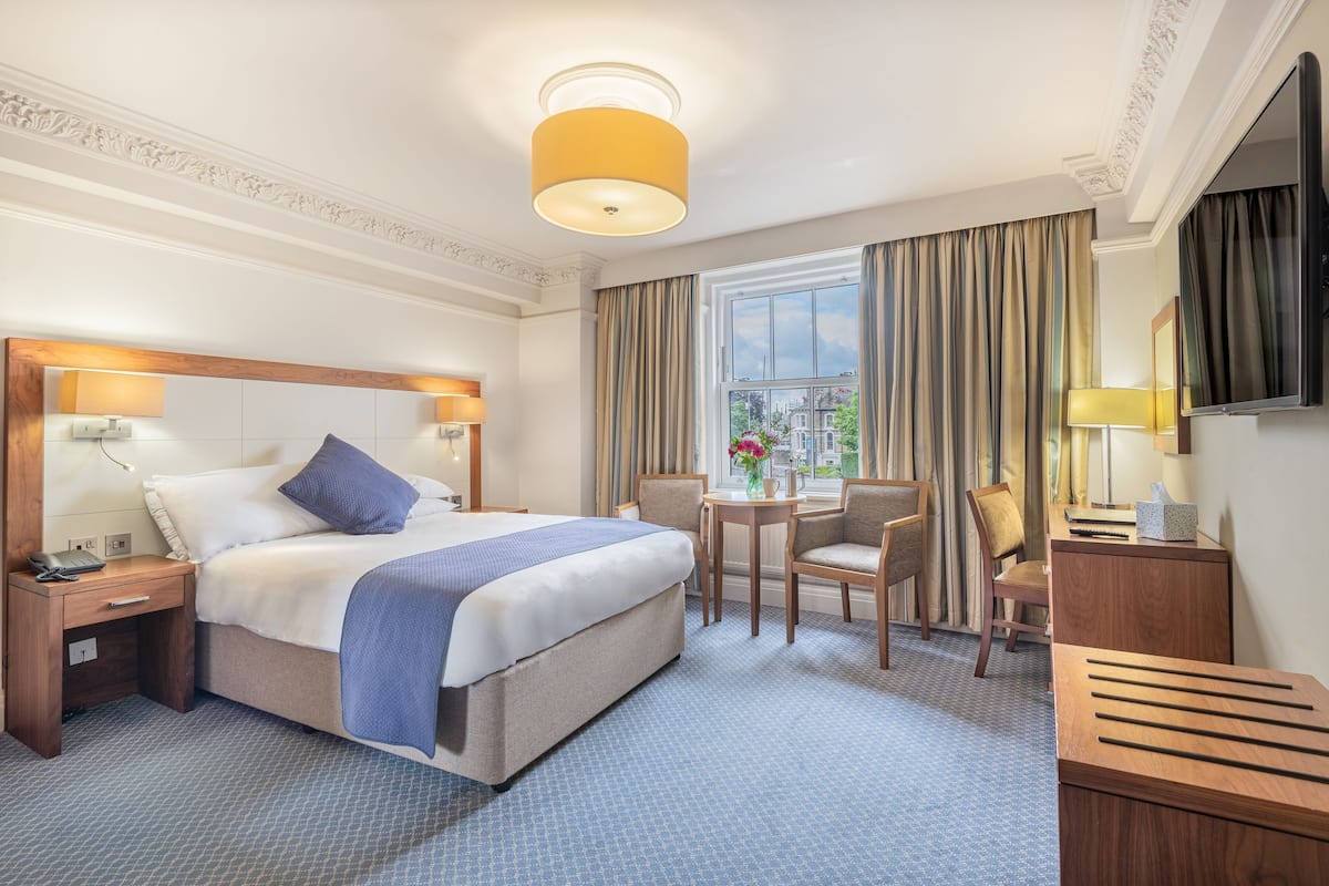 A hotel room with a large bed, a desk, a chair, a small table, and a TV.