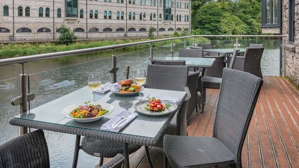 Un espacio de comedor al aire libre con mesas y sillas, barandilla de vidrio y vistas a un río y un edificio al fondo.