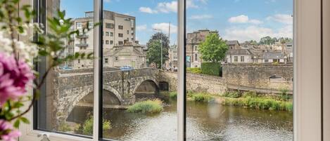 A view through a window of a historic stone bridge over a river, with buildings and greenery in the background.