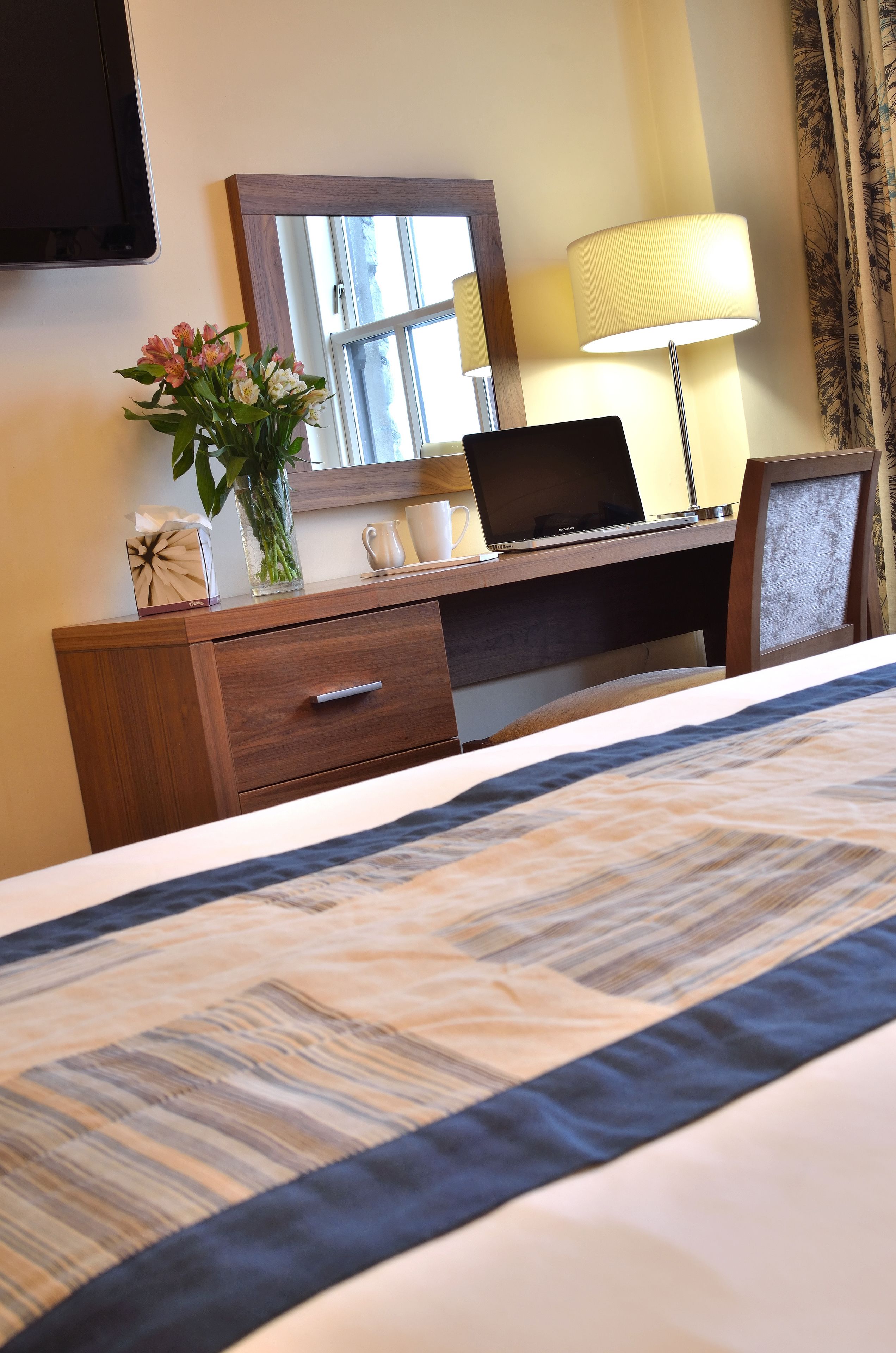 Une chambre d’hôtel avec un lit, un bureau en bois sur lequel se trouve un ordinateur portable, une lampe, un vase avec des fleurs et un miroir.