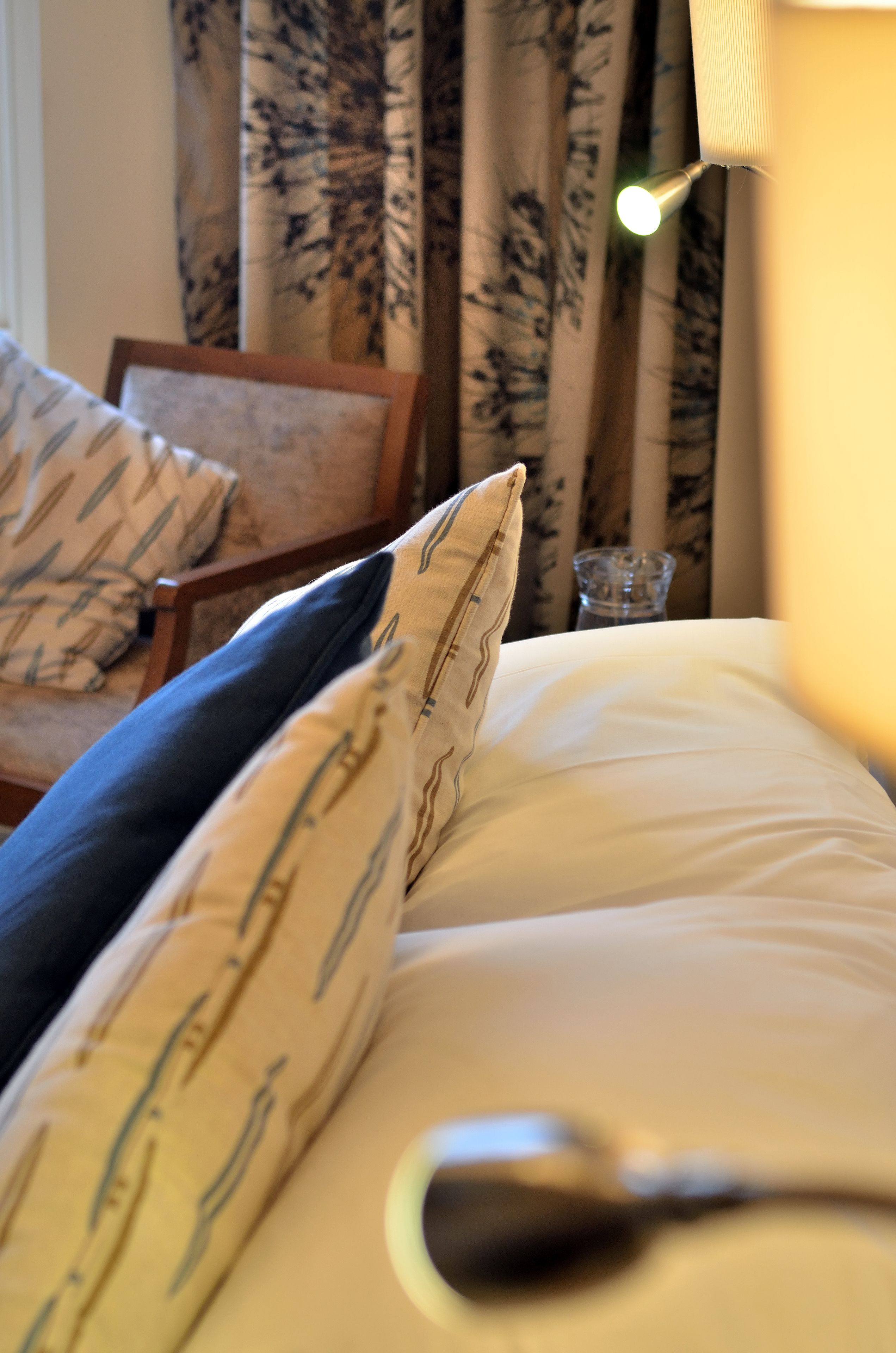 A bed with white sheets and patterned pillows, a bedside lamp, and a chair with a floral curtain in the background.