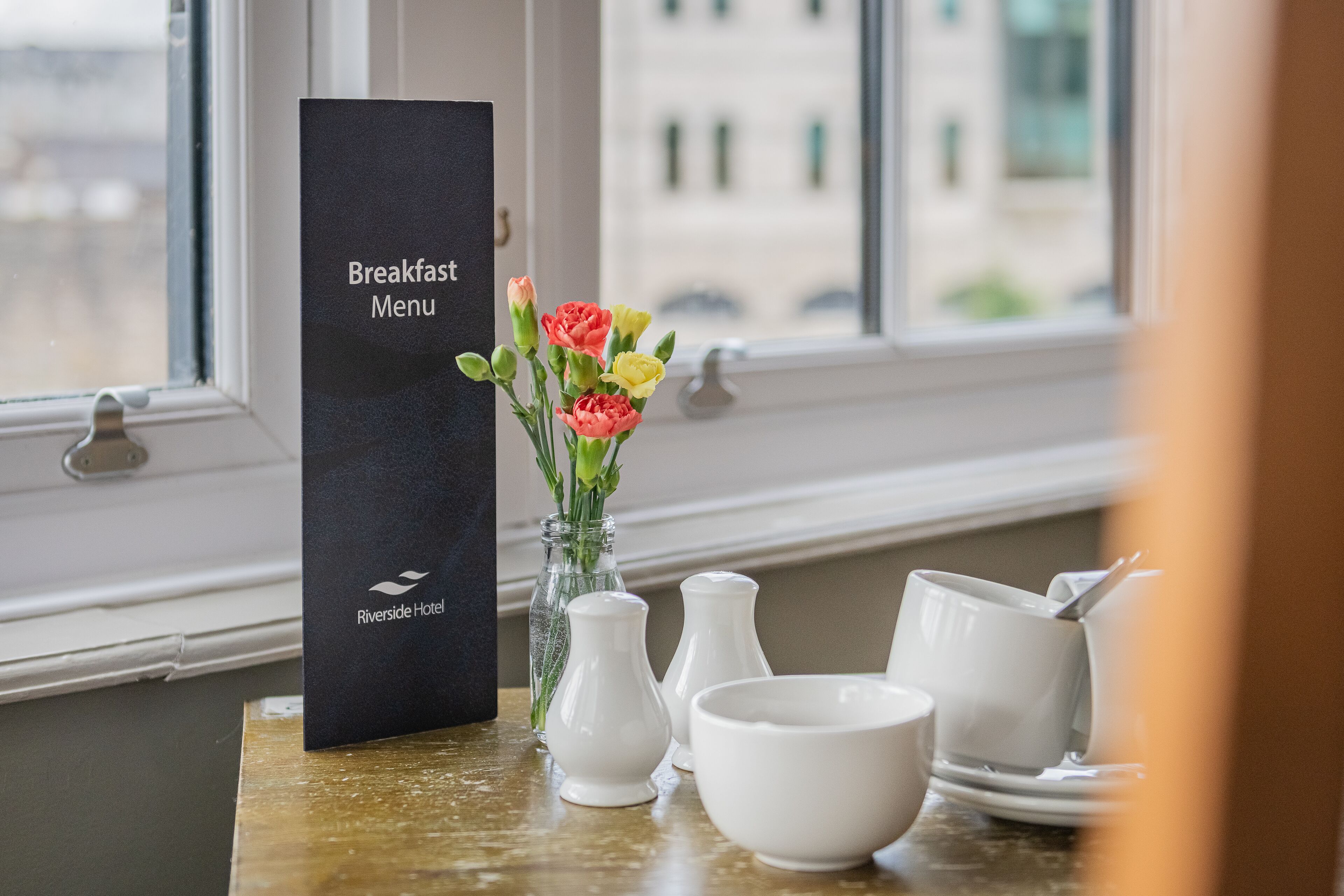 Een menukaartstandaard voor het ontbijt, een vaas met bloemen en wit servies op een tafel bij een raam.