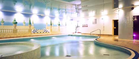 Una piscina interior con un diseño de cuadros en las paredes, un área de descanso con tumbonas y suelo de baldosas.