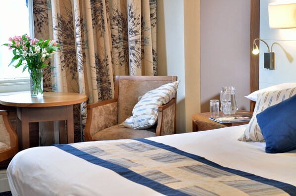Una habitación de hotel con cama, una silla, una mesa con un florero y una lámpara.