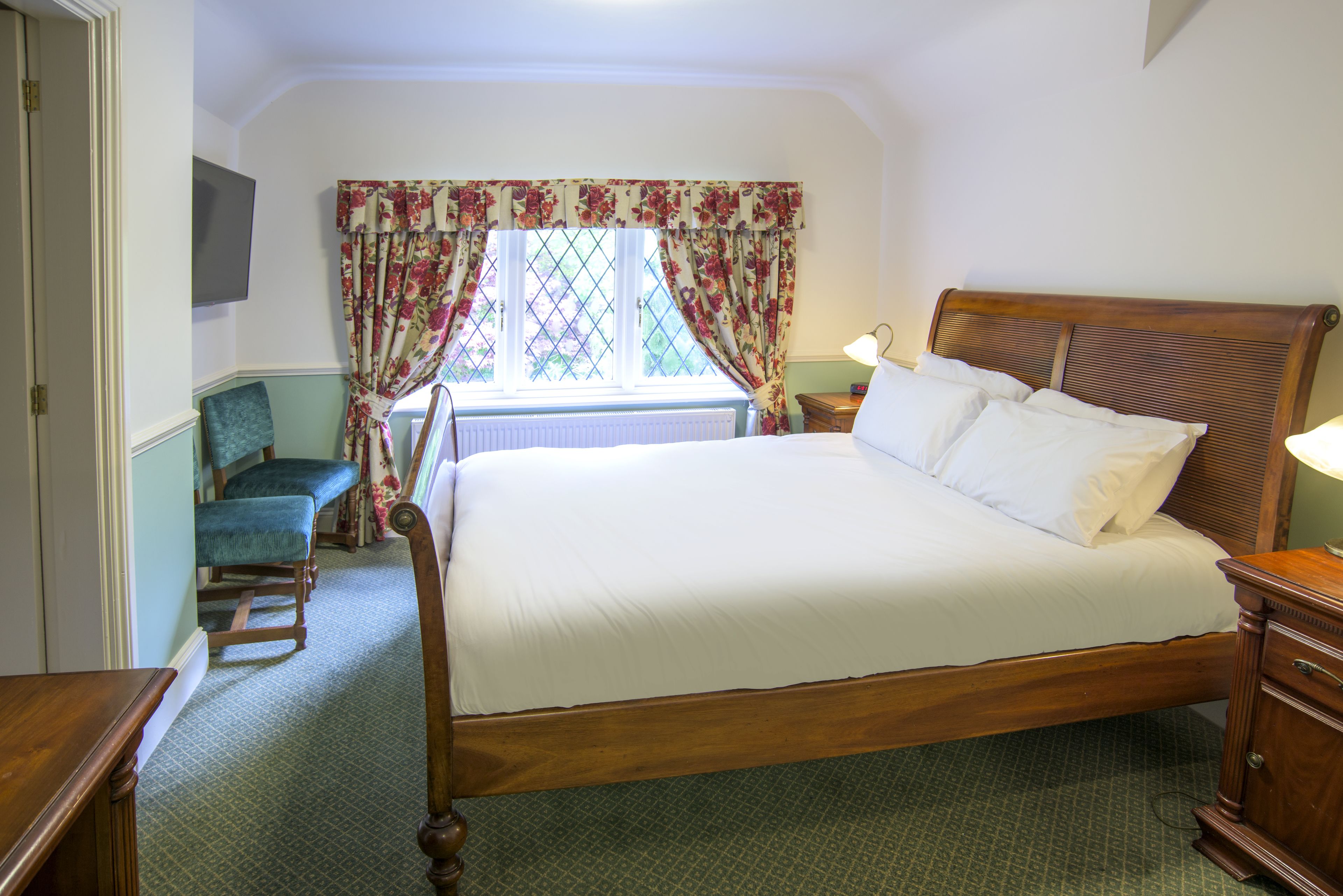 Une chambre à coucher bien rangée, avec un grand lit, une chaise, une table de chevet et une fenêtre ornée de rideaux à motifs floraux.