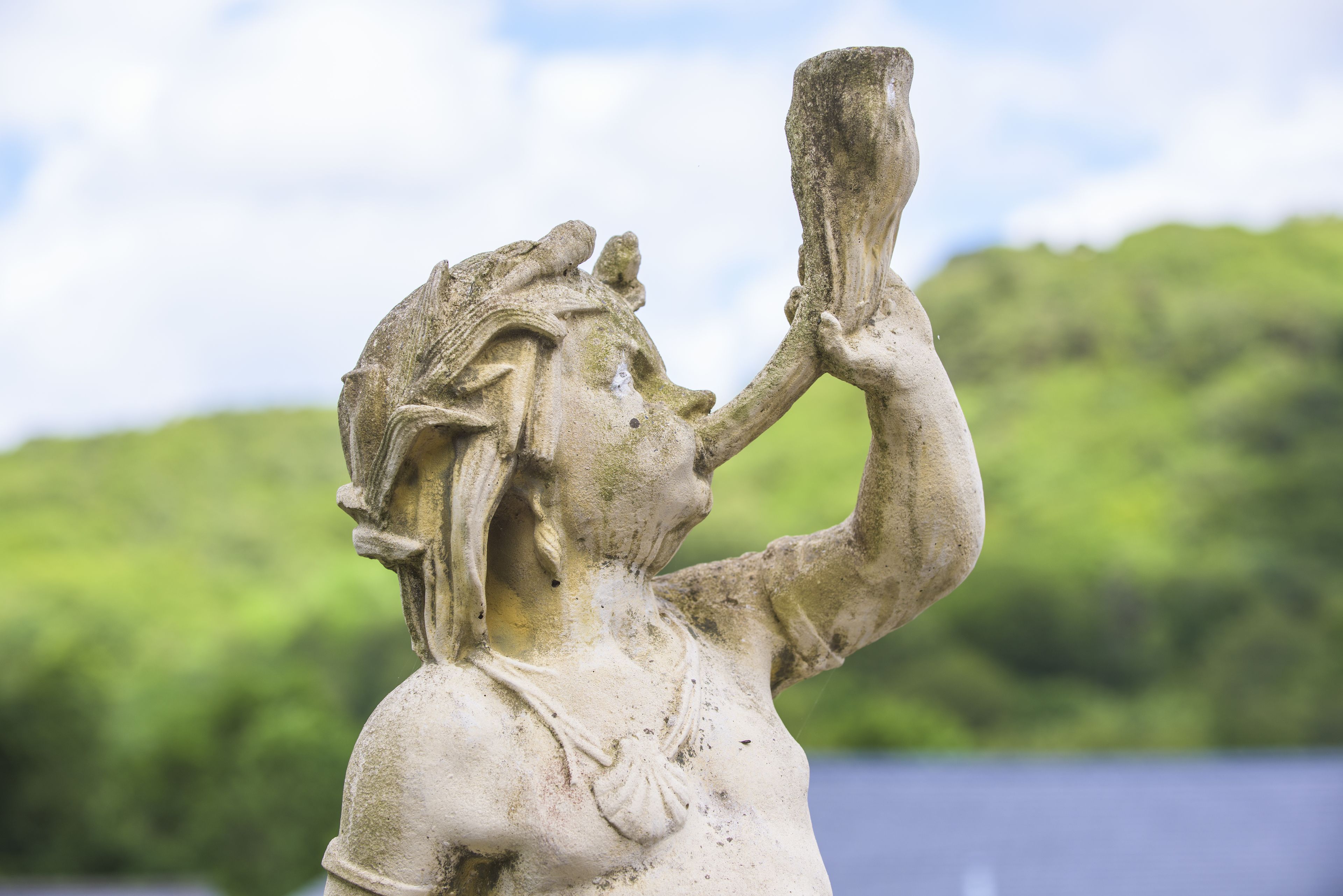 A stone statue of a woman holding a cup, set against a backdrop of greenery and a body of water.