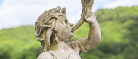 A stone statue of a woman holding a cup, set against a backdrop of greenery and a body of water.