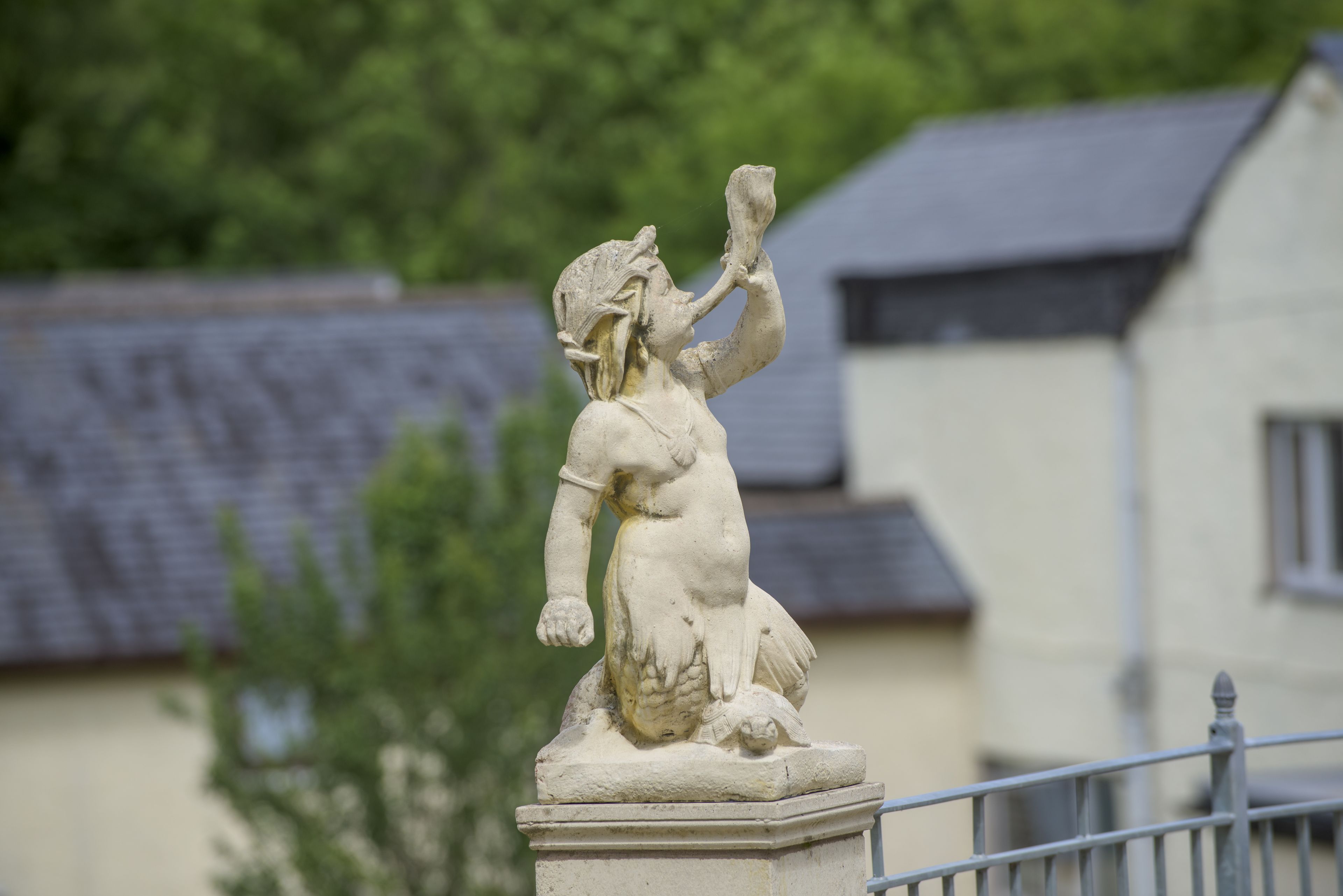 A stone statue of a figure with a raised hand, set on a pedestal, with a building and trees in the background.
