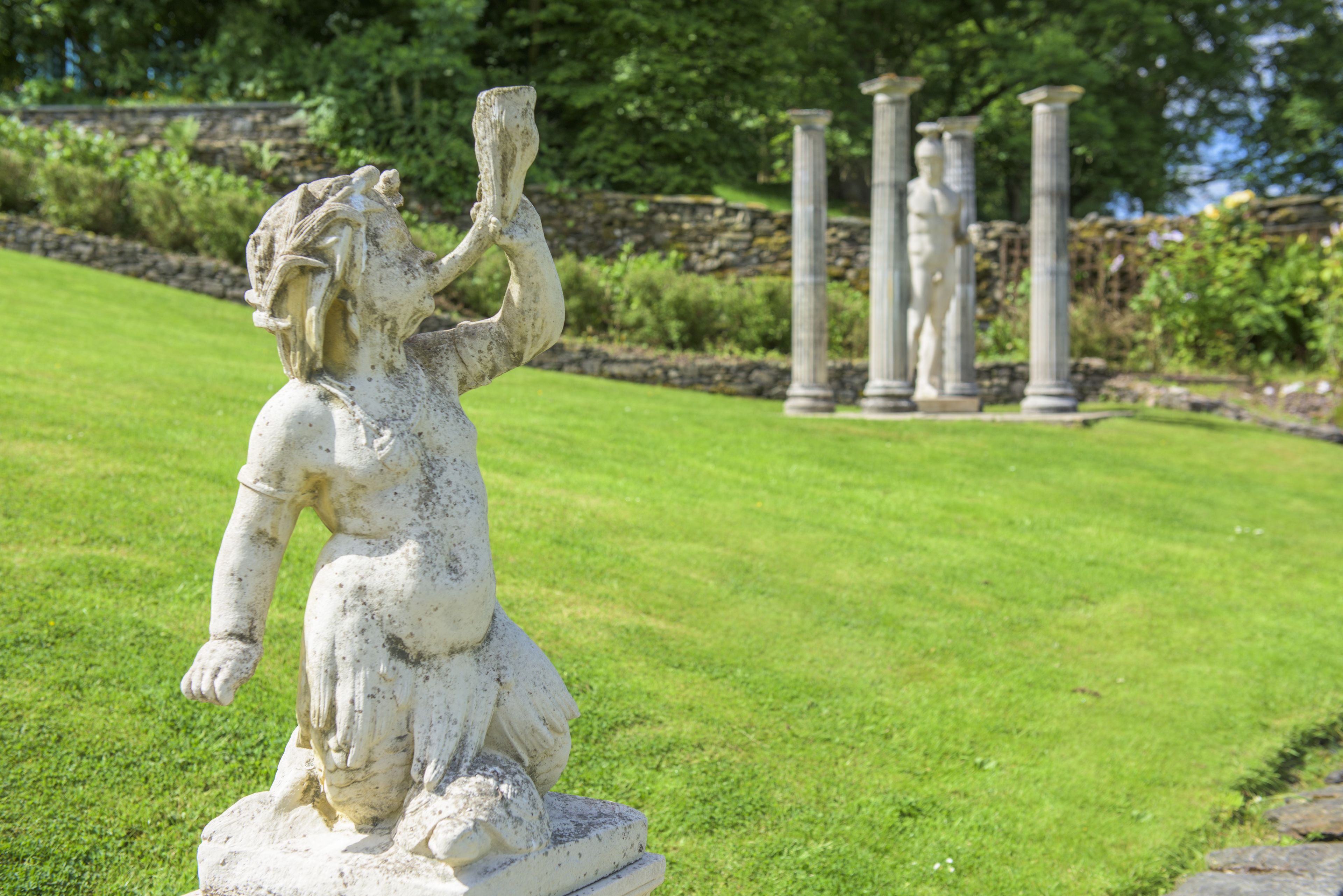 Una estatua de una figura sentada con un brazo levantado, rodeada de un césped cuidado y columnas clásicas al fondo.