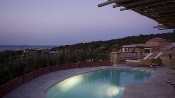 A swimming pool with a curved edge, surrounded by a low stone wall and lounge chairs. In the background, there is a view of the sea and a building with a tiled roof.