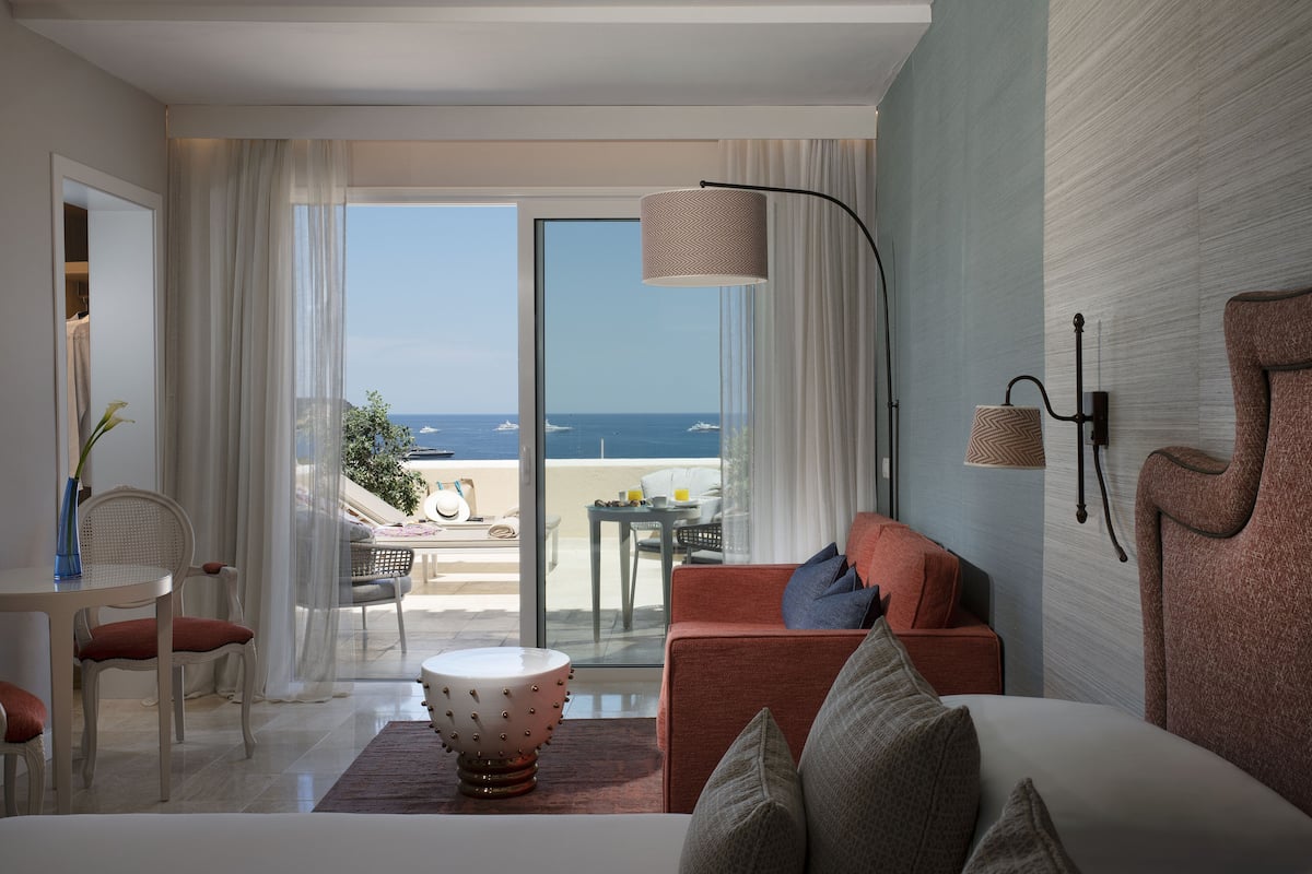 A modern hotel room with a large sliding glass door leading to a balcony with a view of the sea. There is a sofa, a dining table with chairs, and a decorative lamp.