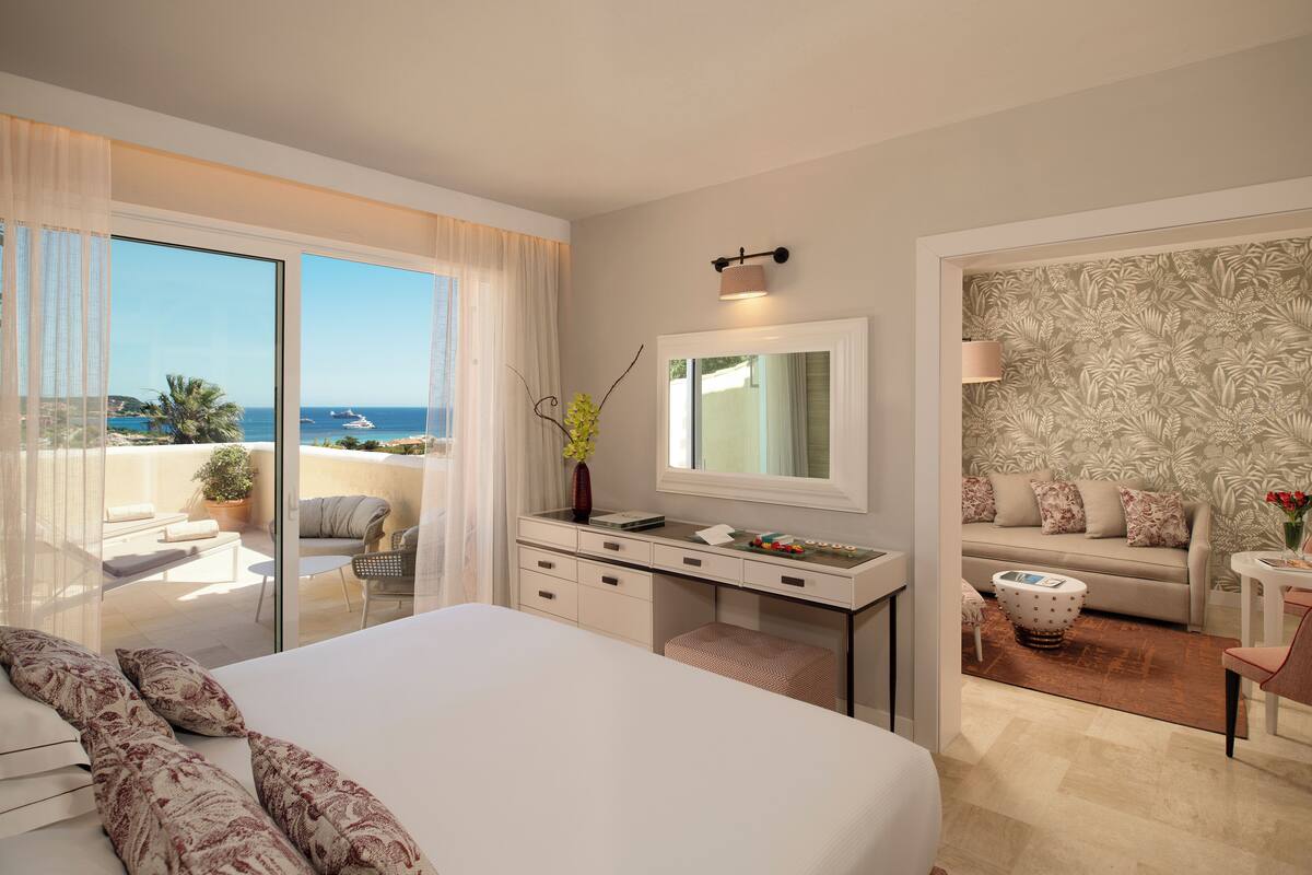 A modern hotel room with a large bed, a desk with a mirror, a balcony with a view of the sea, and a patterned wall.